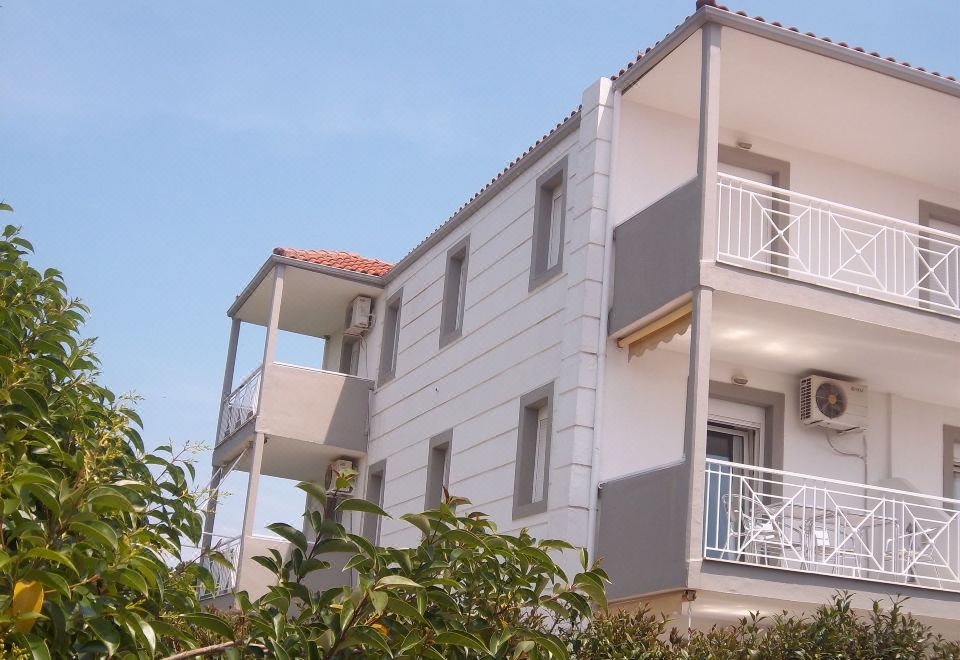 a three - story building with balconies and a red roof is surrounded by greenery , under a clear blue sky at Milos Apartments