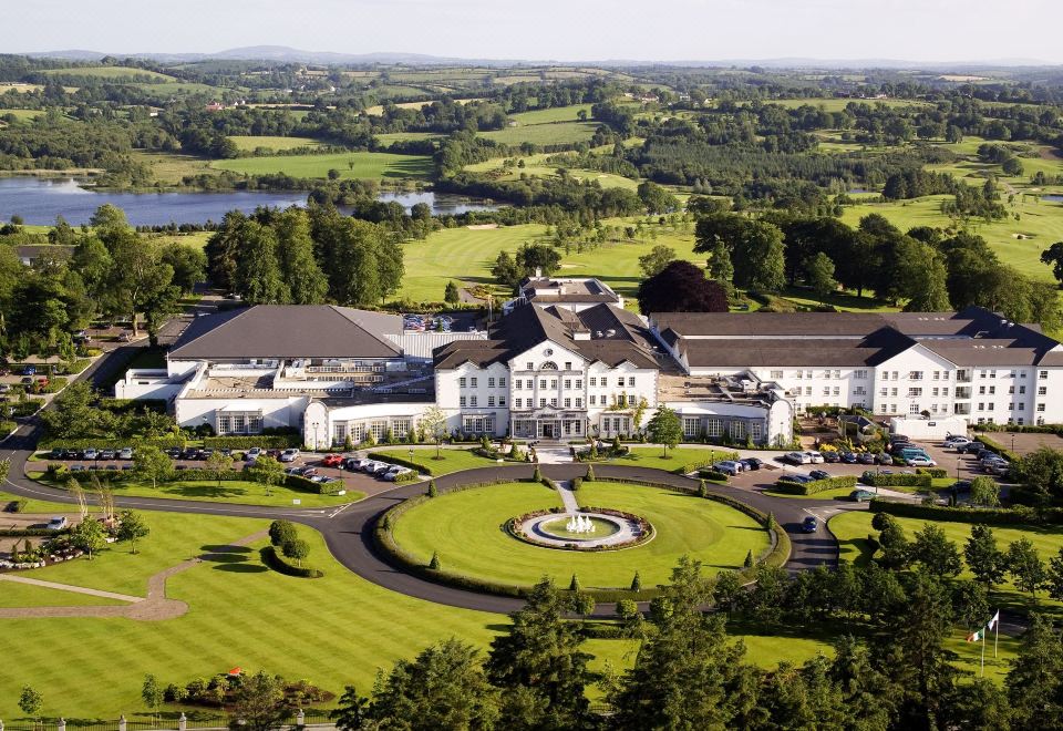 aerial view of a large white building surrounded by green grass and trees , with a lake in the background at Slieve Russell Hotel