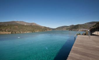 a wooden pier extending into a calm blue - green lake surrounded by mountains under a clear blue sky at Octant Douro