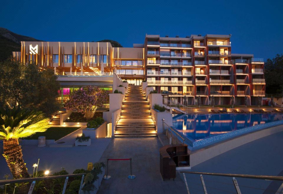 a nighttime view of a hotel with multiple floors , balconies , and a pool , surrounded by trees at Maestral Resort & Casino