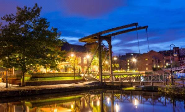 a suspension bridge spanning a body of water , illuminated at night , with a city skyline visible in the background at The Ascott
