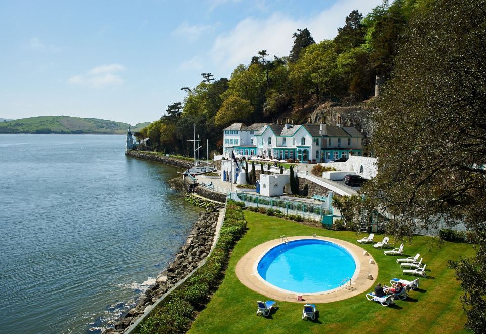 a beautiful waterfront property with a swimming pool , umbrellas , and lounge chairs , surrounded by lush greenery at Portmeirion Village & Castell Deudraeth