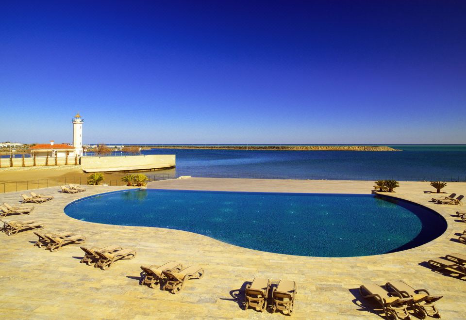 a large outdoor pool surrounded by lounge chairs and a lighthouse , with the ocean in the background at Sheraton Grand Samsun Hotel