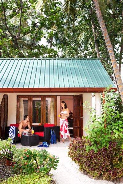 a man and a woman standing in front of a small wooden house , surrounded by lush greenery at Vilamendhoo Island Resort & Spa