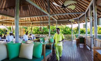a man wearing a green suit is walking through an outdoor dining area , surrounded by wooden tables and chairs at Six Senses Laamu