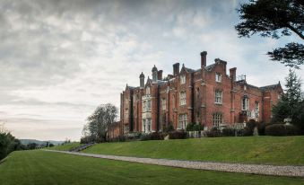 a large red brick building with multiple windows and chimneys , situated on a grassy hillside at De Vere Latimer Estate