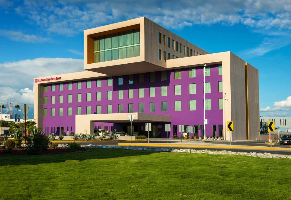 a modern building with a purple facade and large windows is surrounded by green grass and trees at Hilton Garden Inn Monterrey Airport