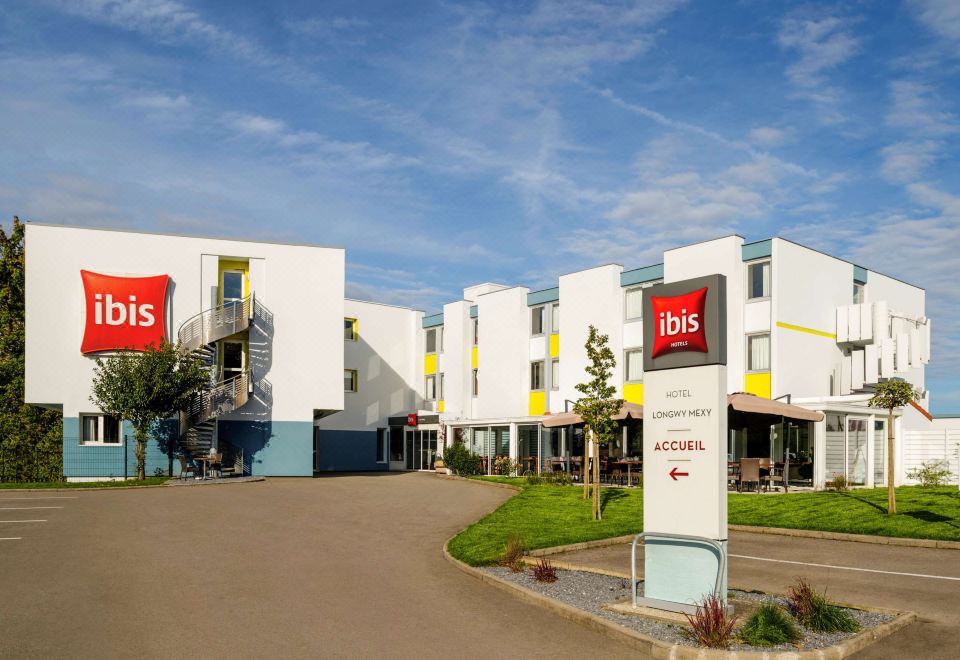 an exterior view of a hotel with a parking lot in front , surrounded by trees and grass at Ibis Longwy Mexy