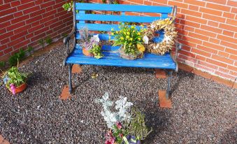 a blue wooden bench placed in a garden , surrounded by potted plants and flowers , with a brick wall in the background at Gila