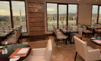 a modern dining room with white chairs and tables , large windows , and a view of the city at Pullman Zamzam Madina