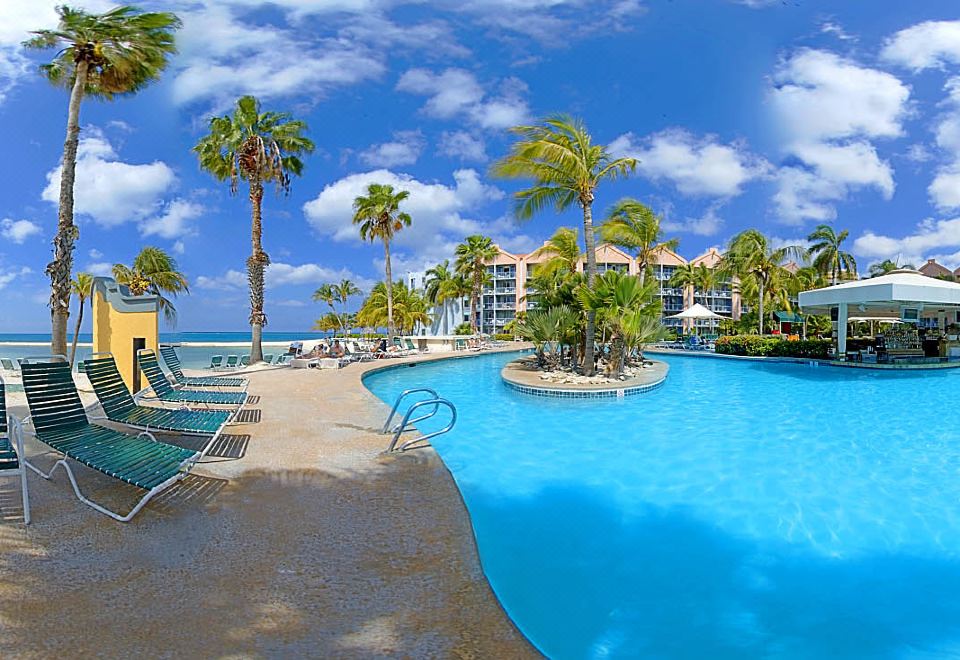 a large outdoor pool surrounded by palm trees and chairs , with a beach in the background at Renaissance Wind Creek Aruba Resort