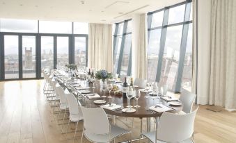 a long dining table is set with white chairs and wine glasses in a room with large windows at Hope Street Hotel