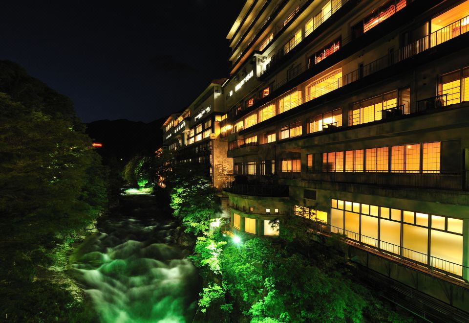 a building with green lights illuminating the night sky , and a river flowing nearby with trees on either side at Zazan Minakami