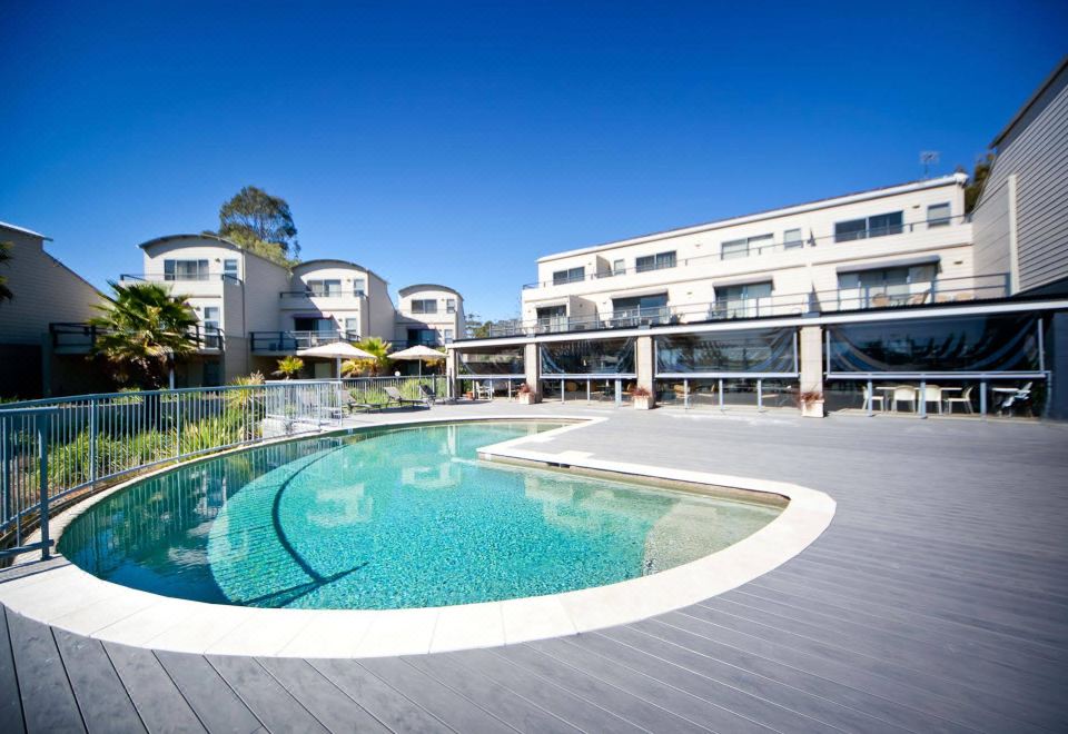 a large , curved pool is surrounded by a modern building with balconies and umbrellas on the ground floor at Corrigans Cove