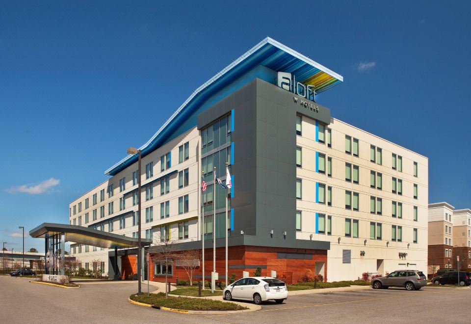 a large hotel with a blue and white facade , surrounded by trees and a parking lot at Aloft Chesapeake