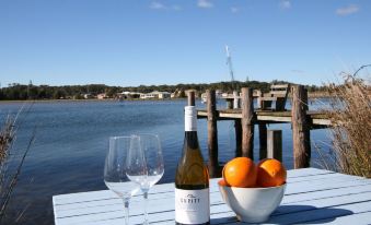 a table with a bottle of wine , two wine glasses , and a bowl of oranges is set up on a dock overlooking the water at Lake Edge Holiday Units