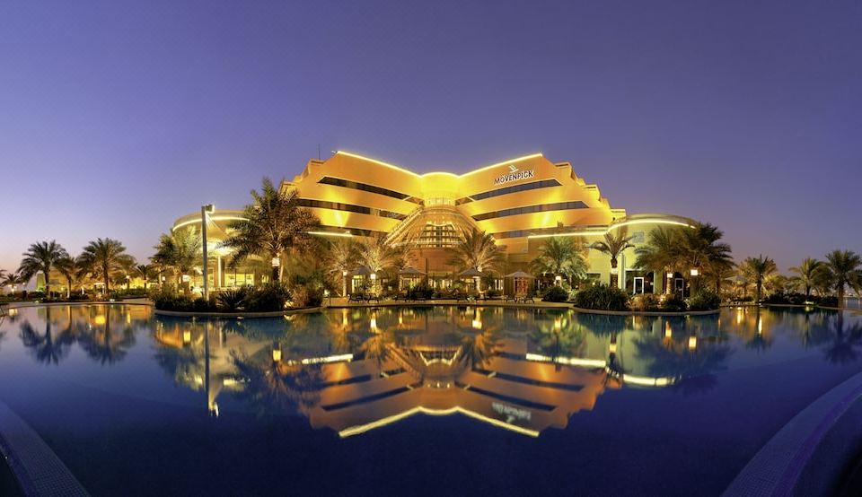 a large hotel with a pool in front of it , surrounded by palm trees and lit up at night at Movenpick Hotel Bahrain