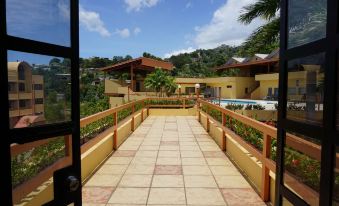 a long , open walkway with a wooden railing and tile flooring leads to a building with red roofs at Ambassador Hotel