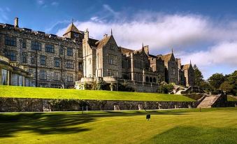 a large stone building with multiple turrets , surrounded by lush green grass and trees , under a blue sky dotted with clouds at Bovey Castle