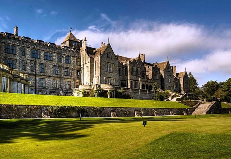 a large , old stone building with multiple turrets , surrounded by green grass and trees , under a blue sky with clouds at Bovey Castle