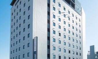 a large white building with a black sign on the side and a clear blue sky in the background at Dormy Inn Mishima