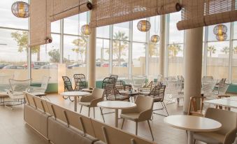 a modern , well - lit restaurant with large windows offering views of the ocean and palm trees at B&B Hotel Barcelona Viladecans
