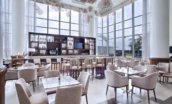 a modern restaurant with large windows , white chairs , and a bar area filled with bottles at Guyana Marriott Hotel Georgetown