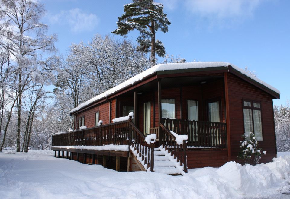 a wooden house surrounded by snow - covered trees , with stairs leading up to the entrance of the building at Loch Kinord Hotel