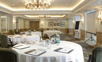 a well - lit conference room with multiple round tables , white tablecloths , and chandeliers , set for a meeting or event at Macdonald Compleat Angler