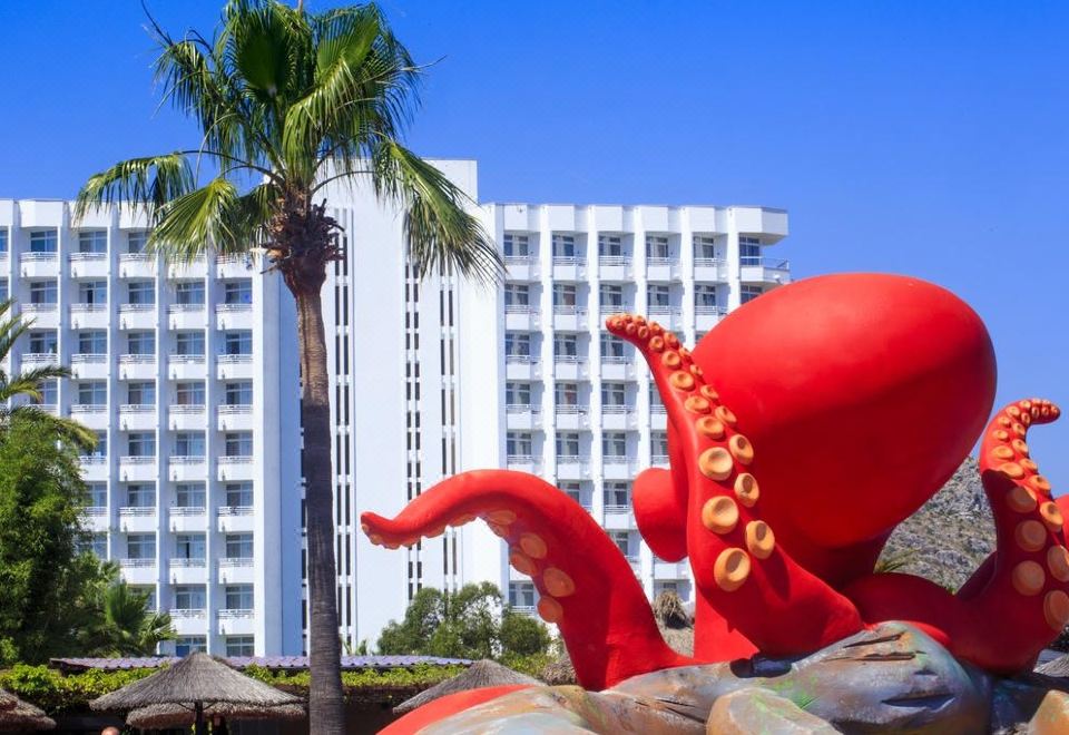 a red octopus sculpture in front of a tall building , surrounded by palm trees and blue sky at Club Mac Alcudia