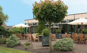 an outdoor dining area with tables , chairs , and umbrellas , surrounded by greenery and a building at Fletcher Hotel Steenwijk