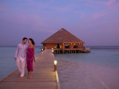 a man and a woman are walking on a wooden pier , leading to a wooden house on stilts at Vilamendhoo Island Resort & Spa