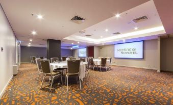 a large conference room with multiple tables and chairs , a projector screen displaying the meeting by novotel logo , and an orange and gray carpet at Novotel Canberra