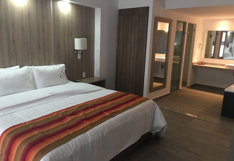 a well - lit hotel room with a large bed , white sheets , and a wooden headboard , as well as a bathroom next to it at Hangar Inn Guadalajara Aeropuerto