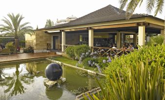 a modern building with a covered terrace , people sitting at tables , and greenery around a small pond at Protur Biomar Sensatori Resort