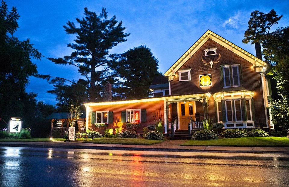 a well - lit house with a sign in front of it , surrounded by trees and a rainy street at The Inn