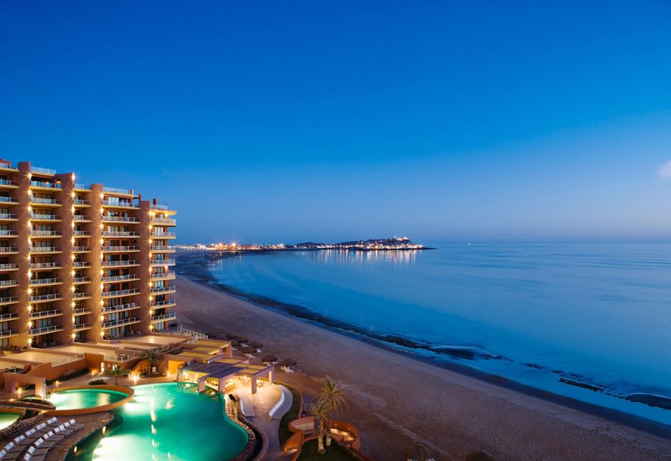 a beach scene with a building on the left side and a pool in the foreground at Las Palomas Beach & Golf Resort