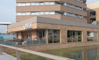 a brick building with a large glass window , located near a body of water and surrounded by trees at Leonardo Hotel Papendrecht