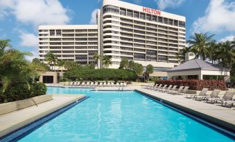 a large hotel with a swimming pool and sun loungers , surrounded by palm trees and blue sky at Hilton Miami Airport Blue Lagoon