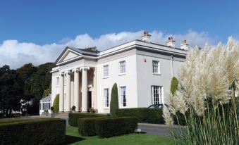 a large white building with a red roof and columns is surrounded by green grass and bushes at Best Western Lamphey Court Hotel