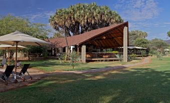 a large wooden structure with a brown roof is surrounded by lush greenery and has a covered area in the foreground at Ashnil Samburu Camp