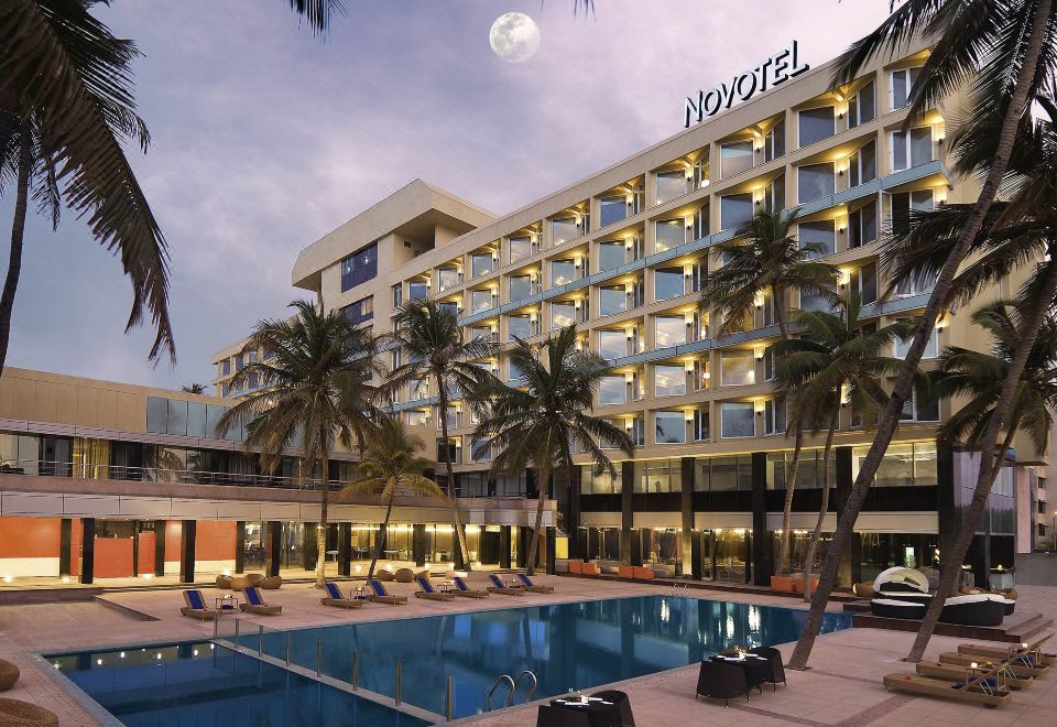 a large hotel building with a pool and palm trees , under a cloudy sky with the moon visible in the background at Novotel Mumbai Juhu Beach