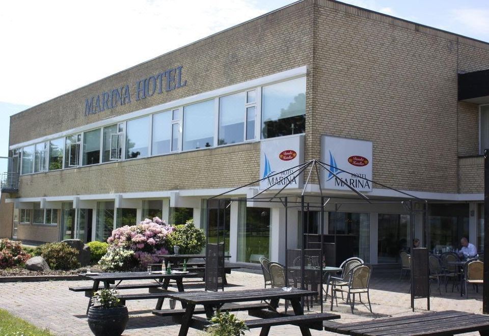 "a brick building with a large sign that says "" marina hotel "" above the entrance , surrounded by a dining area with tables and chairs" at Hotel Marina