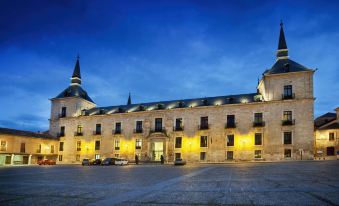 a large building with a pointed roof is illuminated by yellow lights at night , and several cars parked in front of it at Parador de Lerma