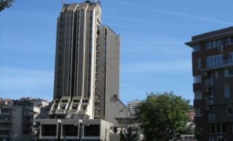 a tall building with a gray facade and a clock on the side is surrounded by trees at Zlatibor Hotel