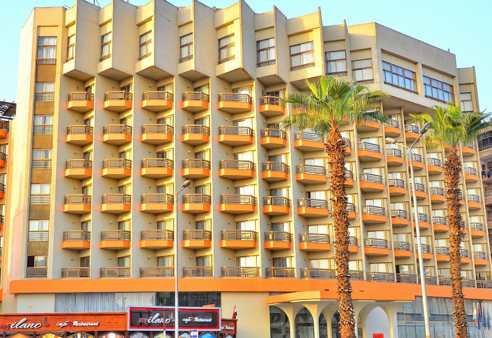 a modern , multi - story building with balconies and orange accents , surrounded by palm trees and advertisements in arabic at Aracan Pyramids Hotel