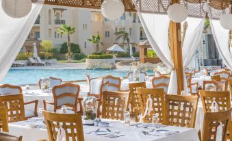 a poolside restaurant with white tablecloths , chairs , and tables set up under a wooden structure , overlooking a pool area at Kempinski Hotel Soma Bay