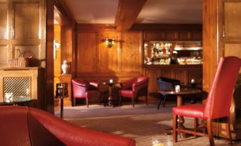 a cozy wooden bar area with red chairs , a piano , and wine glasses on the table at Macdonald Compleat Angler