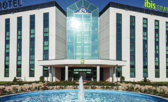 a modern building with a large glass facade , surrounded by lush greenery and a blue fountain at Ibis Styles Milano Est Settala