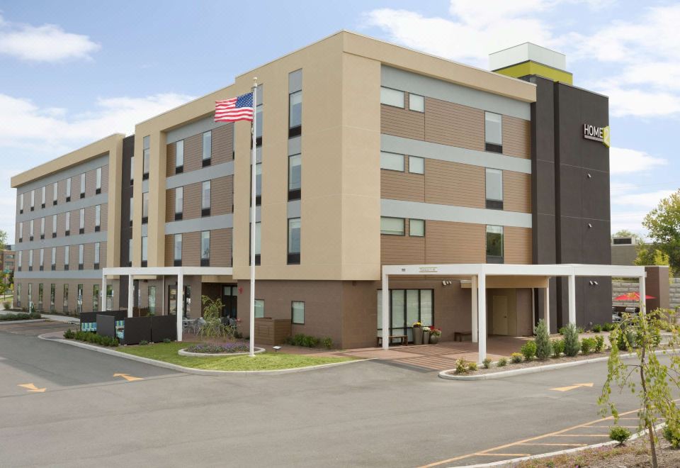 a modern hotel building with a tan facade and white columns , featuring an american flag on the left side at Home2 Suites by Hilton Rochester Henrietta